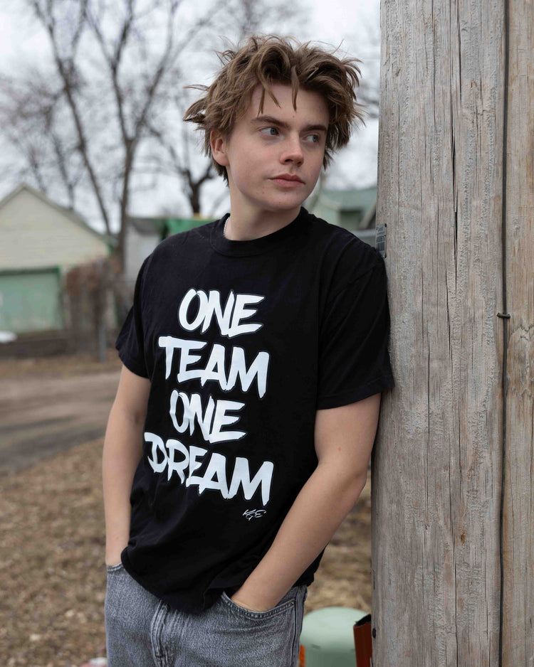 Young male model wearing a black “One Team One Dream” Comfort Colors T-shirt while posing outdoors against a wooden post.